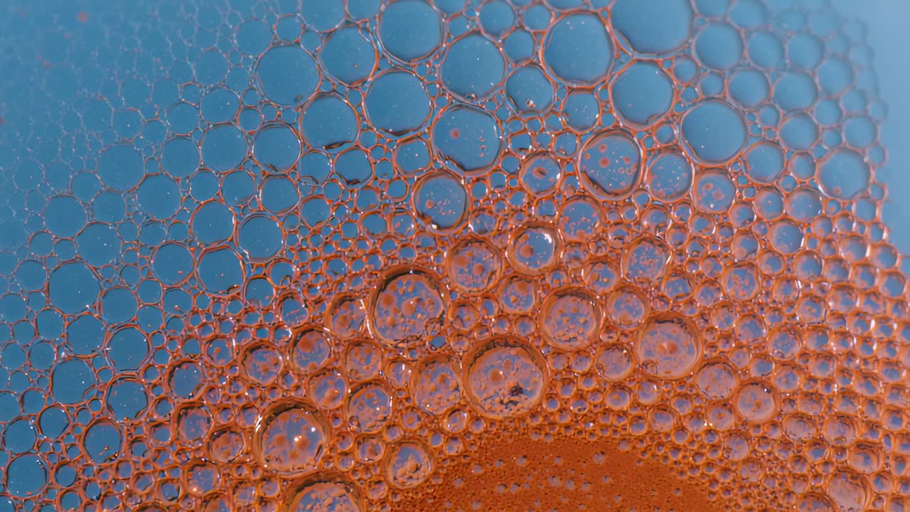 Ammasso di schiuma arancione che avanza spostandosi verso l'alto, con un movimento fluido che fa unire le bolle in un primo piano blu.