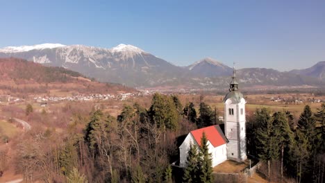 drone flying from the church away from it with large mountains in the background. karavanke alps in the background with snow on the mountains. village nestled in the alps of slovenia.