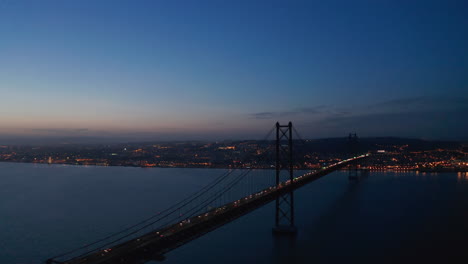 Vista-Aérea-Nocturna-Del-Puente-25-De-Abril-Que-Conecta-Lisboa-Y-Almada.-Faros-De-Automóviles-En-El-Puente-De-La-Carretera-Atirantado-Sobre-El-Río-Tegus.-Drone-Volando-Hacia-Adelante.-Lisboa,-Capital-De-Portugal.