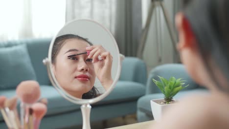 reflection in a mirror beautiful young asian woman combing long black eyelashes with mascara brush and smiling while applying makeup at home