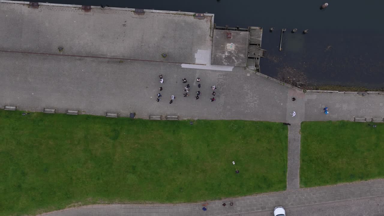 Aerial top-down view of a group of people gathered on a waterfront promenade beside a dock and green lawns. Scenic drone footage capturing human activity and public space