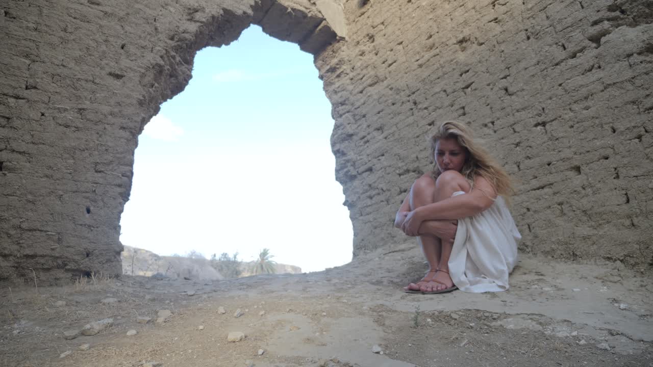 A woman in a white dress huddles alone under a deserted ruin