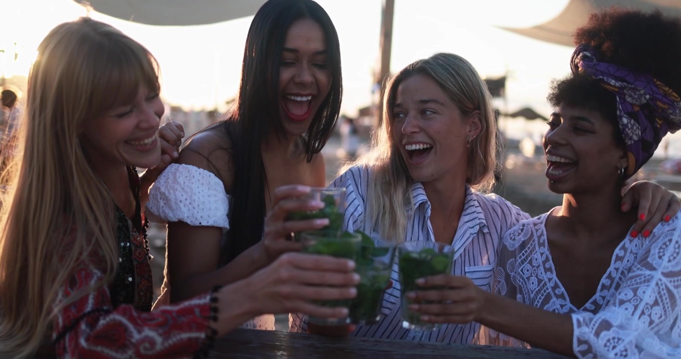 Glade unge kvinner skåler med cocktails på en strandbar ved solnedgang