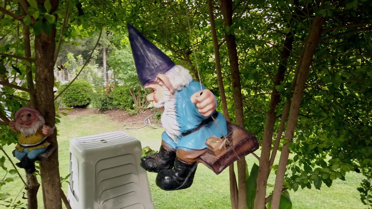 Garden gnome decoration seated on a wooden swing among trees and green foliage, with a blue hat, white beard, and colorful clothing visible in a garden in Mexico.