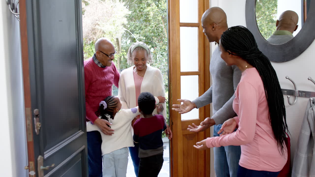 Happy african american parents, son and daughter welcoming grandparents at front door, slow motion