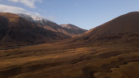 Aerial---golden,-wavy-plains-in-front-of-snow-capped-hills-in-Mackenzie,-New-Zealand