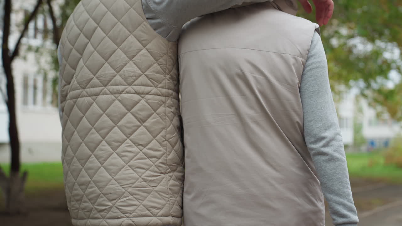 Back view of two people walking closely in matching light quilted summer jackets with one placing hand gently on other's back, outdoor scene with soft background trees and buildings partially visible