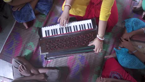 married indian teacher teaches music with harmonium at rural classroom with students gathered around, unrecognizable, top down shot