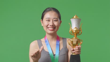 close up of asian woman with a gold medal showing gold trophy to camera, smiling, and pointing herself being proud winning as the first winner on green screen background in the studio