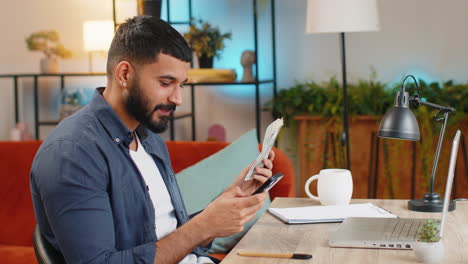 Rich-Indian-man-freelancer-using-smartphone-counting-money-cash-calculate-earnings-at-home-office