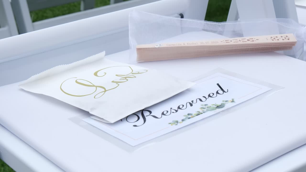 Elegant close-up of a white wedding chair marked reserved with an envelope and a hand fan. Set on lush green grass, perfect for capturing summer wedding details and decor