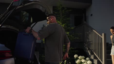 happy guy in sunglasses together with his girlfriend loading bags into the trunk of a car while leaving the hotel in summer