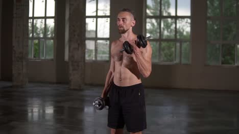 Shirtless-muscular-man-doing-workout-with-dumbbells-and-sweat-on-his-body.-Man-shirtless-with-sweaty-torso-exercising-in-empty-loft-studio.-Slow-motion