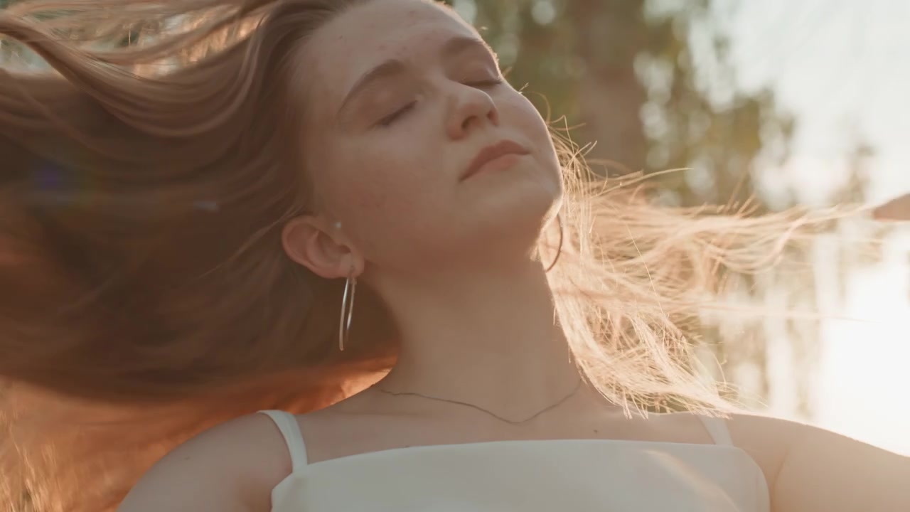 Una donna con i capelli lunghi si passa le mani tra i capelli in un ambiente esterno, indossando un top bianco e orecchini a cerchio d'argento. Il suo abbigliamento di colore chiaro contrasta delicatamente con la vegetazione sfocata degli alberi.