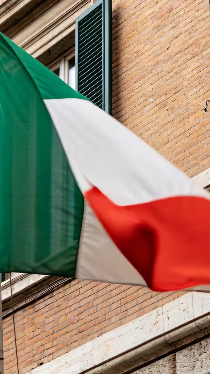 Video verticale: Bandiera tricolore che sventola su un'asta sopra una facciata di mattoni mentre le raffiche spingono il tessuto.