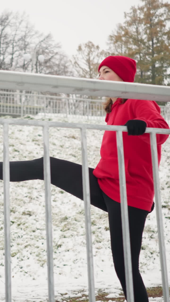 woman exercising outdoors during winter stretching leg back and forth with hand wearing black gloves against snowy ground foggy atmosphere distant trees background breath visible motion detail