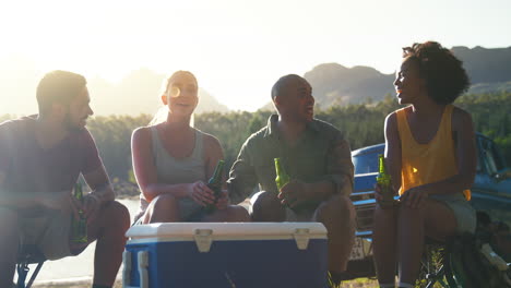 grupo de amigos con mochilas en una camioneta en un viaje por carretera bebiendo cerveza de un refrigerador en el lago