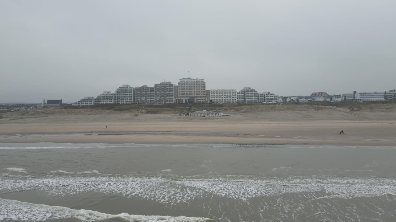 Waves gently roll onto the beach with a Dutch coastal city skyline in the background on a grey morning.