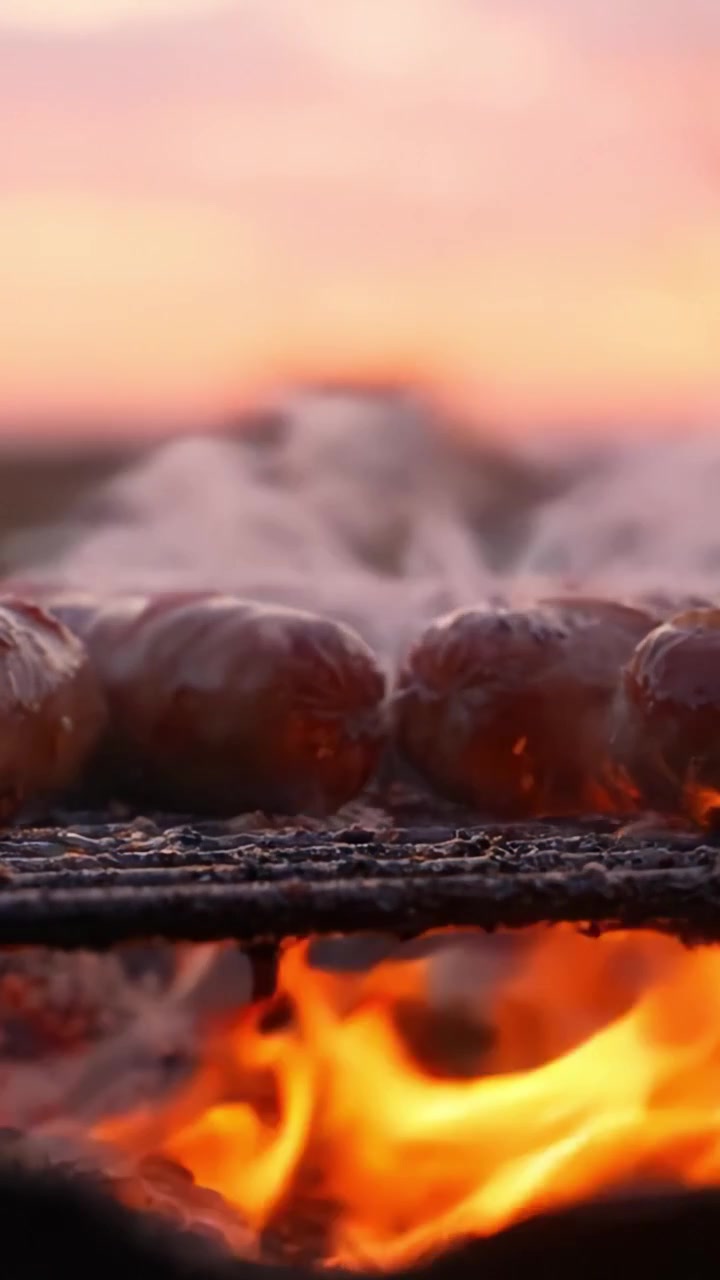 Salsicce sfrigolanti su una griglia con fiamme vivaci e fumo che creano una deliziosa esperienza di barbecue al tramonto, mettendo in mostra una delizia culinaria e un'atmosfera di cucina all'aperto.