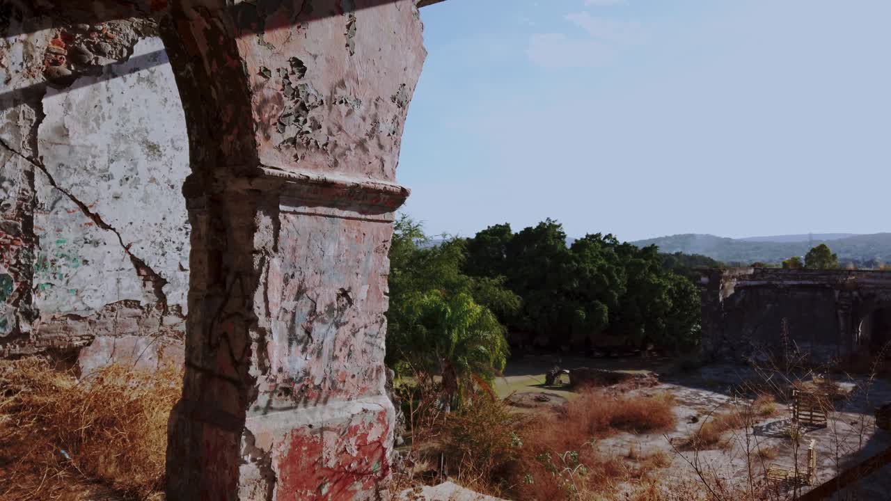 Interior de hacienda abandonada con paredes rotas y pilares arqueados, luz solar proyectando sombras en el suelo, hierba seca dentro de la estructura mostrando deterioro, historia y una atmósfera tranquila.