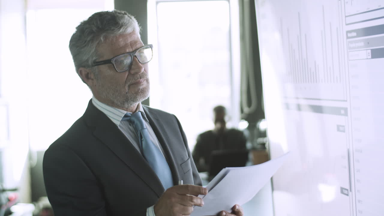 Serious manager in eyeglasses checking documents with data on screen