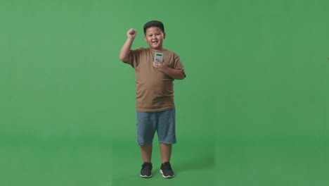 cuerpo lleno de feliz niño asiático celebrando mientras usa teléfono inteligente en pantalla verde en el estudio
