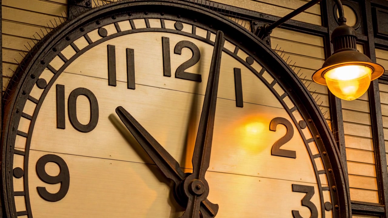 Advancing round wall clock moving minute hand toward hour mark in vintage hall, lamp glow shifting