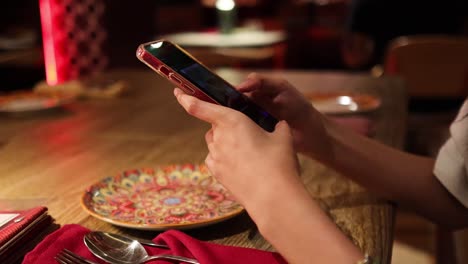 a person interacts with a smartphone at a warmly lit restaurant table, with a colorful plate, utensils, and red napkin visible. static close-up shot