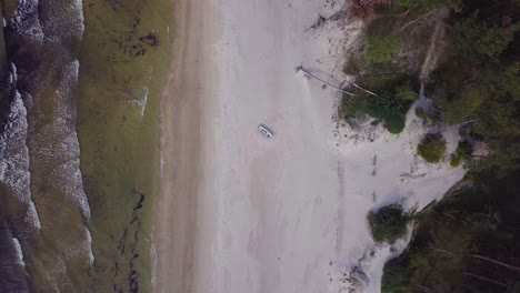 Aerial-birdseye-view-of-Baltic-sea-coast-on-a-sunny-day,-blue-coastal-fisherman-boat,-white-sand-seashore-dunes-damaged-by-waves,-pine-tree-forest,-coastal-erosion,-climate-changes,-wide-drone-shot