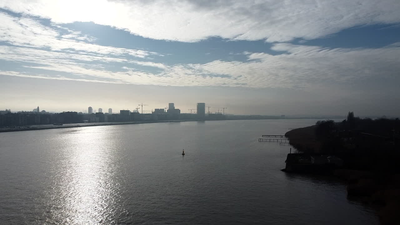 Slow aerial forward shot over scheldt river with sun reflections and buildings in background