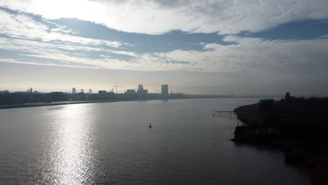 slow aerial forward shot over scheldt river with sun reflections and buildings in background