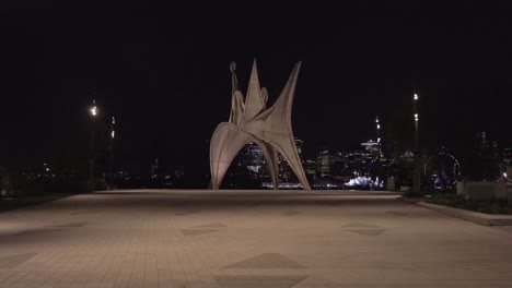 estatua de arte por la noche en el parque jean drapeau en montreal, quebec, con el horizonte iluminado de la ciudad en el fondo