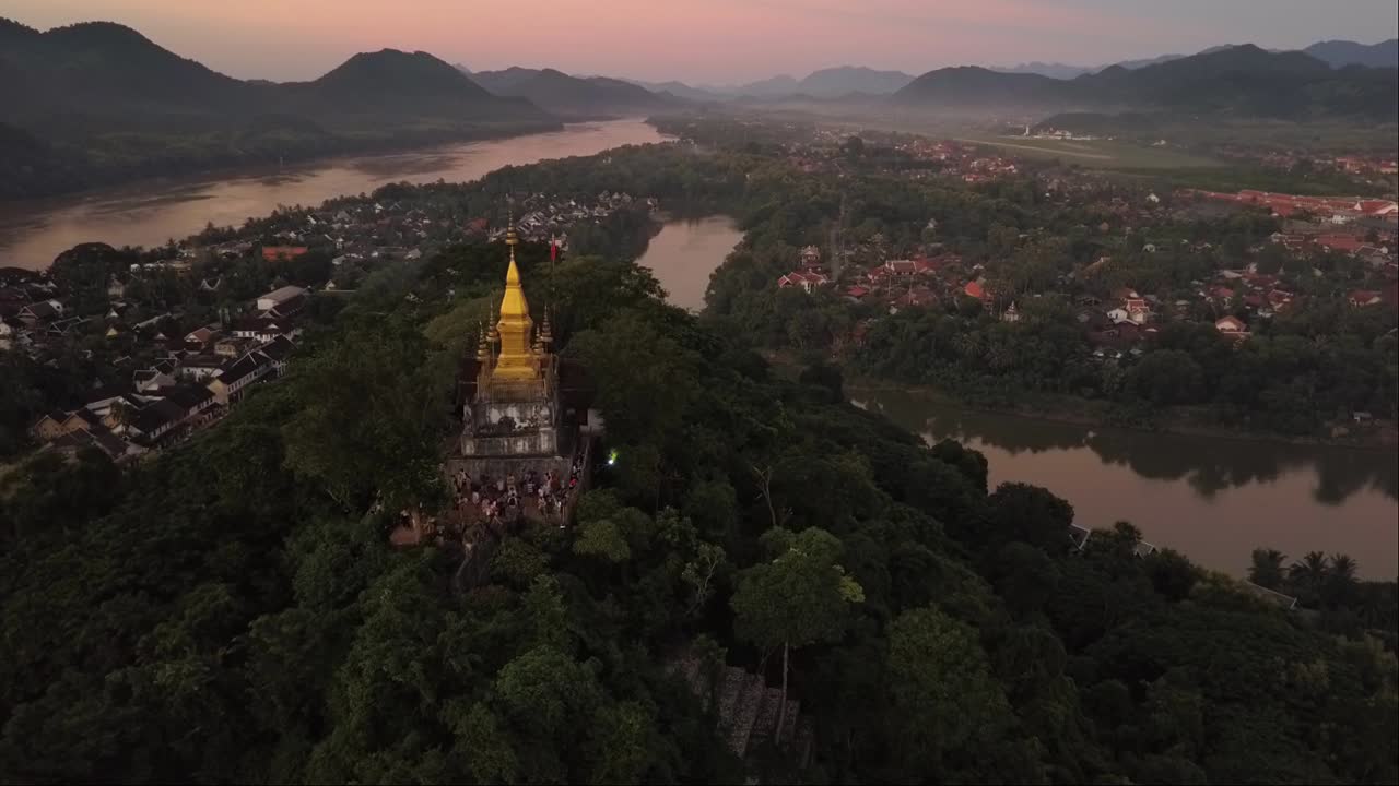 Drone statico che inquadra un tempio solitario situato in cima a un'antica città, circondato da un fiume tranquillo, montagne e dal sole al tramonto.