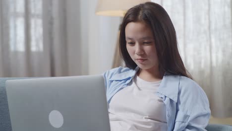 close up of asian teen girl with snacks typing on a laptop while lying on sofa in the living room at home