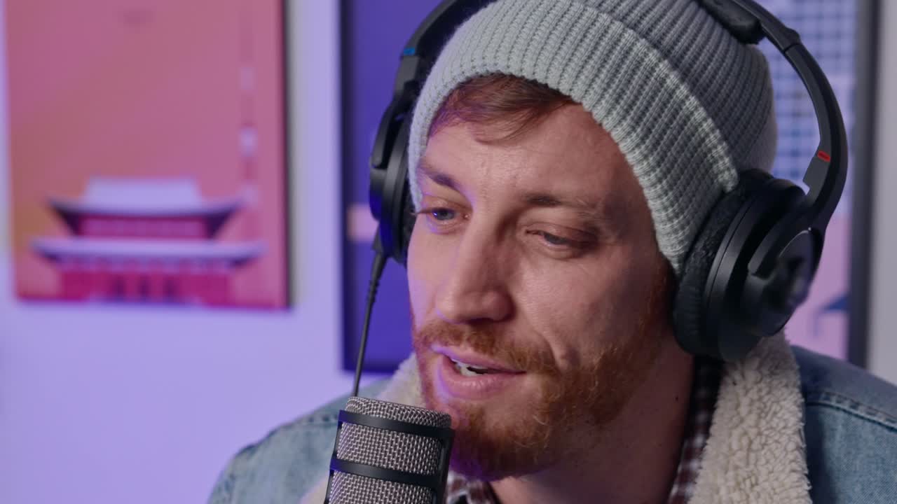 Hipster guy talking to a microphone on live podcast show as guest or host. Man with beanie knit hat and headphones speaks in studio in streaming. Medium shot talking head. Person with beard