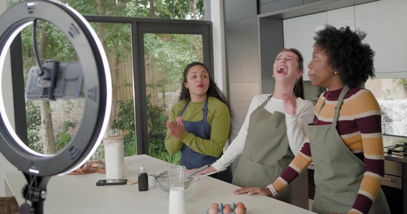 Three adult women friends in aprons filming for ring light waving laughing gesturing at mixing bowl