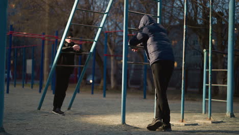 close-up of two individuals resting on an iron bar at an outdoor park, with light casting shadows on the ground and a car passing by in the background