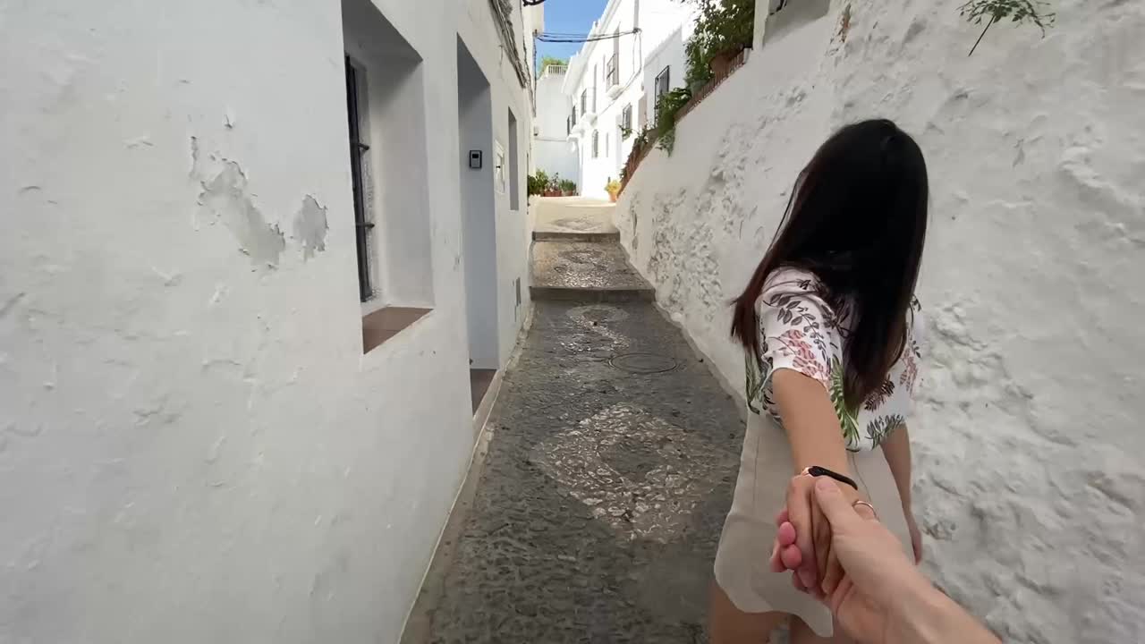 Giovane coppia si tiene per mano passeggiando in uno stretto vicolo bianco a Frigiliana, Malaga. Piante lussureggianti, porte tradizionali in un villaggio andaluso. Filmato di uno stile di vita di viaggio romantico