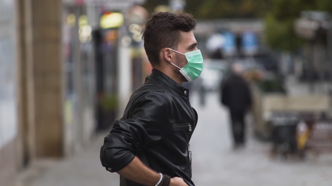 A young man in a leatherette jacket with a modern hairstyle on a busy street, putting on a green protective Covid-19 facemask, putting his hands in his pockets, sideways to the camera, close up 4k