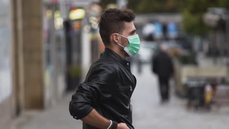 a young man in a leatherette jacket with a modern hairstyle on a busy street, putting on a green protective covid-19 facemask, putting his hands in his pockets, sideways to the camera, close up 4k