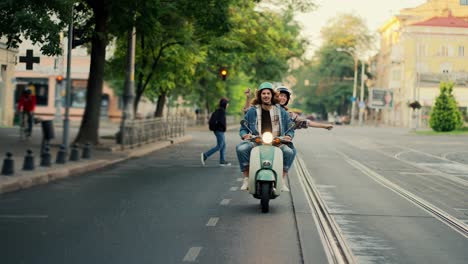 Ein-Glücklicher-Typ-In-Einer-Jeansjacke-Fährt-Mit-Seiner-Freundin,-Die-Ihre-Arme-Bewegt-Und-Tanzt,-Während-Sie-Auf-Einem-Moped-Mit-Eingeschalteten-Scheinwerfern-Auf-Einer-Morgendlichen-Stadtstraße-Im-Sommer-Fährt