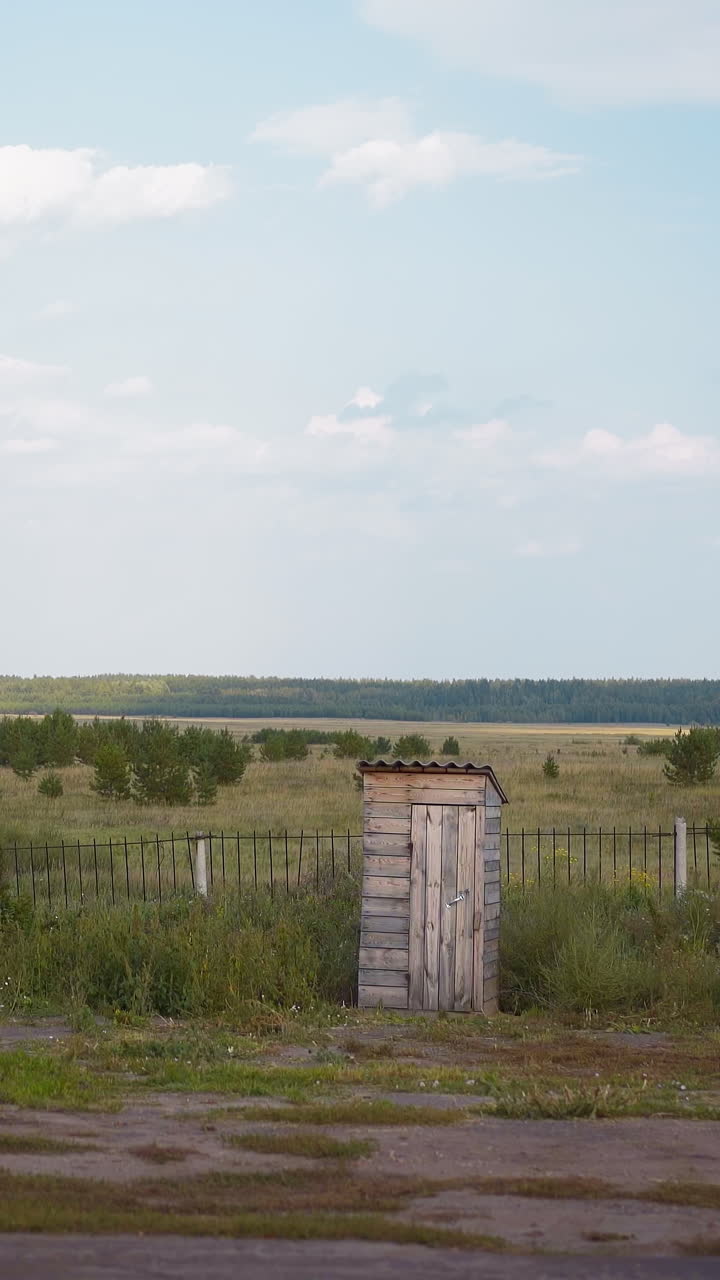 Lonely wooden composting toilet near fence in village house yard. Small shed made of boards for gardening tools keeping. Farmland building against green meadow