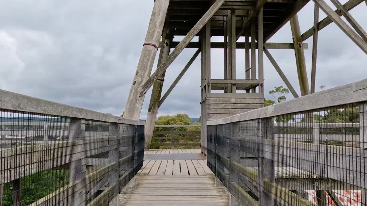 A close-up view of a wooden walkway leading to a rustic observation tower under a cloudy sky.