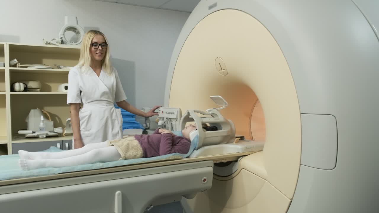 mujer médico radiólogo hace una resonancia magnética del cerebro, cabeza, cuello para el paciente niña pequeña, niña yace en la mesa automática, utilizando equipos modernos, bobina en la cabeza, auriculares