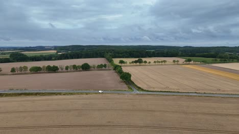 harvested brown wheat fields and driving white car on intersection road. aerial forward wide shot. green hills in rural area of american city. panorama view. cloudy summer day in united states