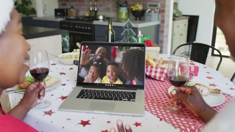 pareja afroamericana con vino usando computadora portátil para la videollamada de navidad con familia feliz en la pantalla