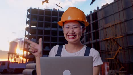 close up de una trabajadora asiática que usa gafas de protección y casco de seguridad usando una computadora portátil y apuntando hacia un lado mientras está de pie en un sitio de construcción