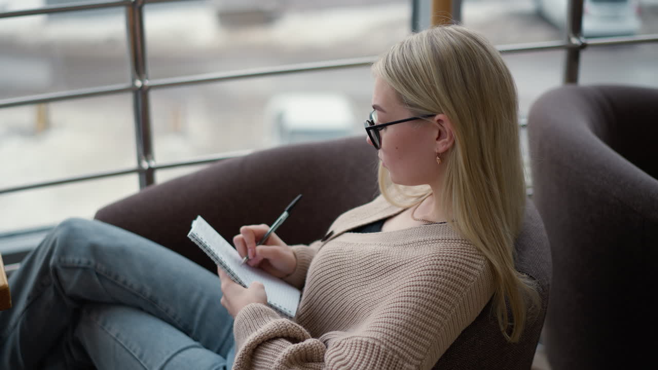 Young woman seated in a cozy indoor space, writing in her notebook with focused expression, capturing a calm and thoughtful moment with pen in hand, looking outside