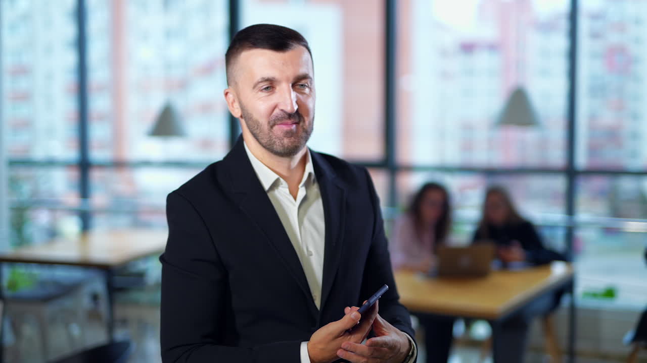 Bearded man receives a call on the phone. Businessman having conversation and gesturing with his hand. Women sitting at the desk at backdrop in blur.