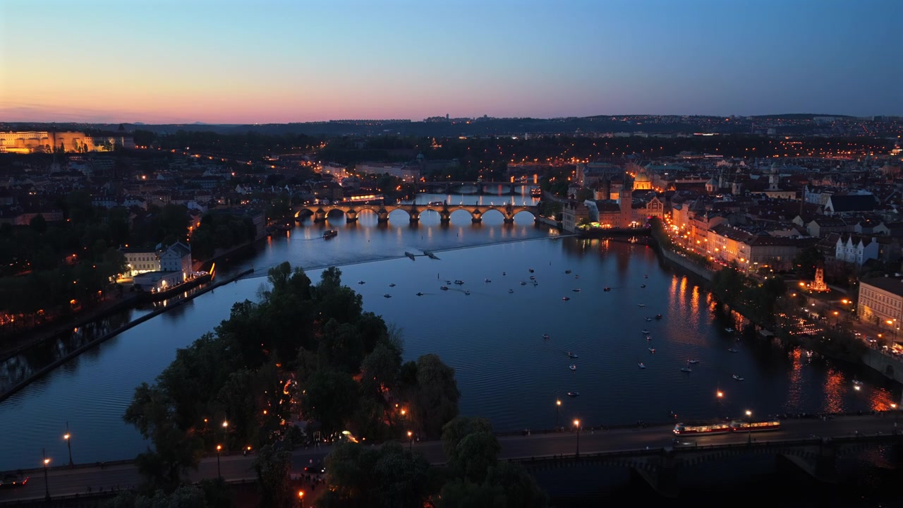 Veduta aerea ad alta quota del Ponte Carlo e del fiume Moldava di notte, con piccole imbarcazioni e la città illuminata sotto un cielo crepuscolare.